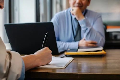 A lady signing a contract,and a man in blue shirt on the other side of the table looks  at her