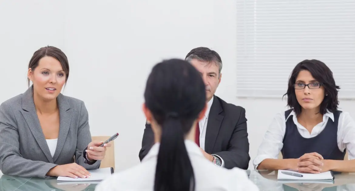Recruitment Agency in qatar-Wings Technical Services A person is seated at a table facing three individuals who appear to be conducting an interview. The person being interviewed has their back to the camera, while the three interviewers are seated across the table, each with documents and a pen in hand. The setting appears to be a professional office environment with white walls and a window with blinds in the background.