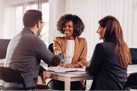 Sure! Here’s a brief alt text for your image:</p>
<p>**Alt text**: Three people are seated at a table in an office setting. Two of them are facing the camera, while the third person, wearing glasses, is facing away. The person in the middle is shaking hands with the person on the left. The table has some documents and a pen on it.