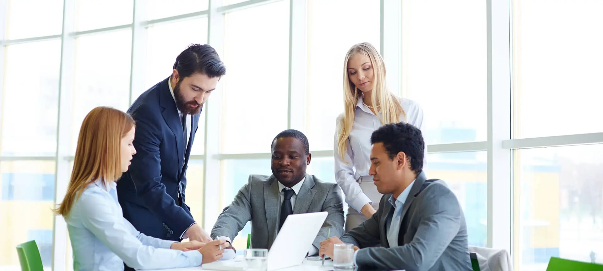 Here's the alt text for the image you provided:</p>
<p>"A group of five people in business attire are gathered around a table in a modern office setting with large windows. One person is standing and appears to be leading the discussion, while the others are seated and engaged, with one person using a laptop."</p>
<p>This alt text should be helpful in describing the image for accessibility purposes. Let me know if there's anything else I can assist you with!