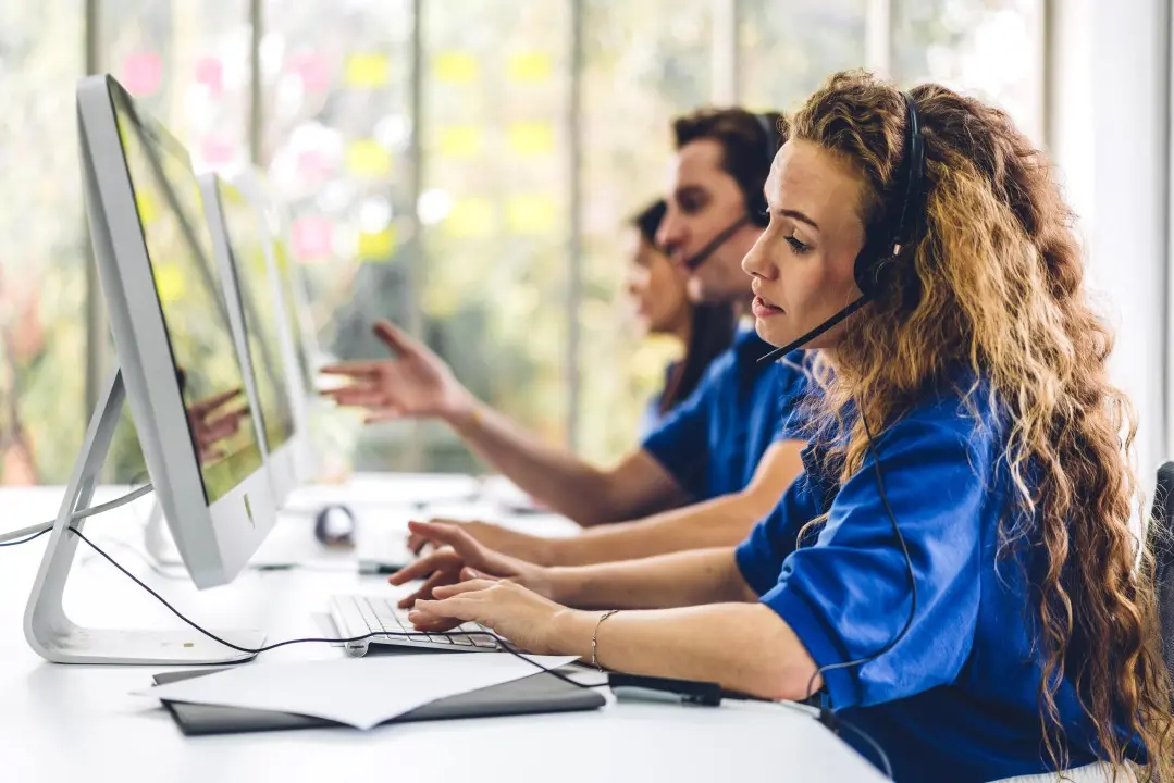 three people in blue uniform sits at a desk wearing headphone engaging in their work
