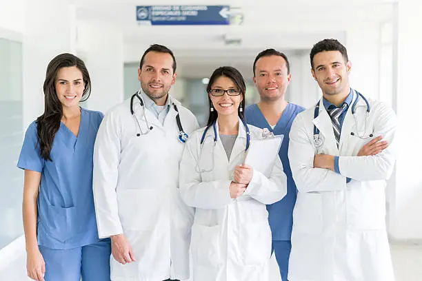 A group of five healthcare professionals standing in a hospital corridor. Three of them are wearing white lab coats with stethoscopes around their necks, and two are wearing blue scrubs. One person in a lab coat is holding a clipboard. There is a sign in the background indicating directions within the hospital.