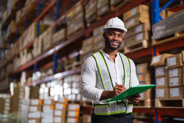A smiling man wearing a hard hat holds a clipboard while standing in a warehouse, showcasing a positive work environment.